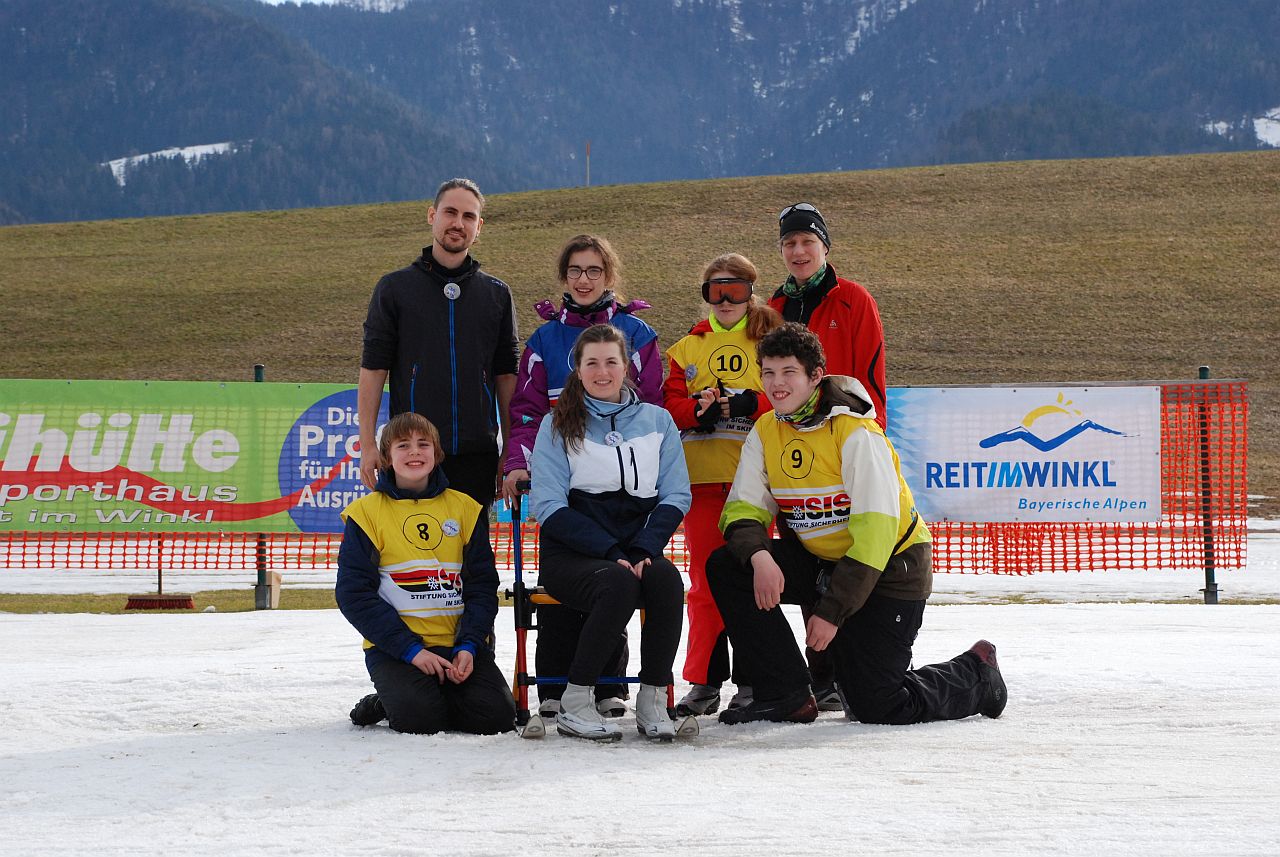 Christopher, Frau Fecke, Julian, Herr Kling, Marija, Lara und Frau Gauch stehen im Schnee vor einer Absperrung zur Loipe. Im Hintergrund sieht man einen grünen Hügel und verblaute Berge.