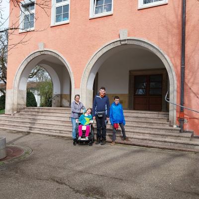 Im Hintergrund ist eine Kirche , vor der Treppe ist eine Rollstuhlfahrerin , zwei stehende Schüler und eine Betreuerin 