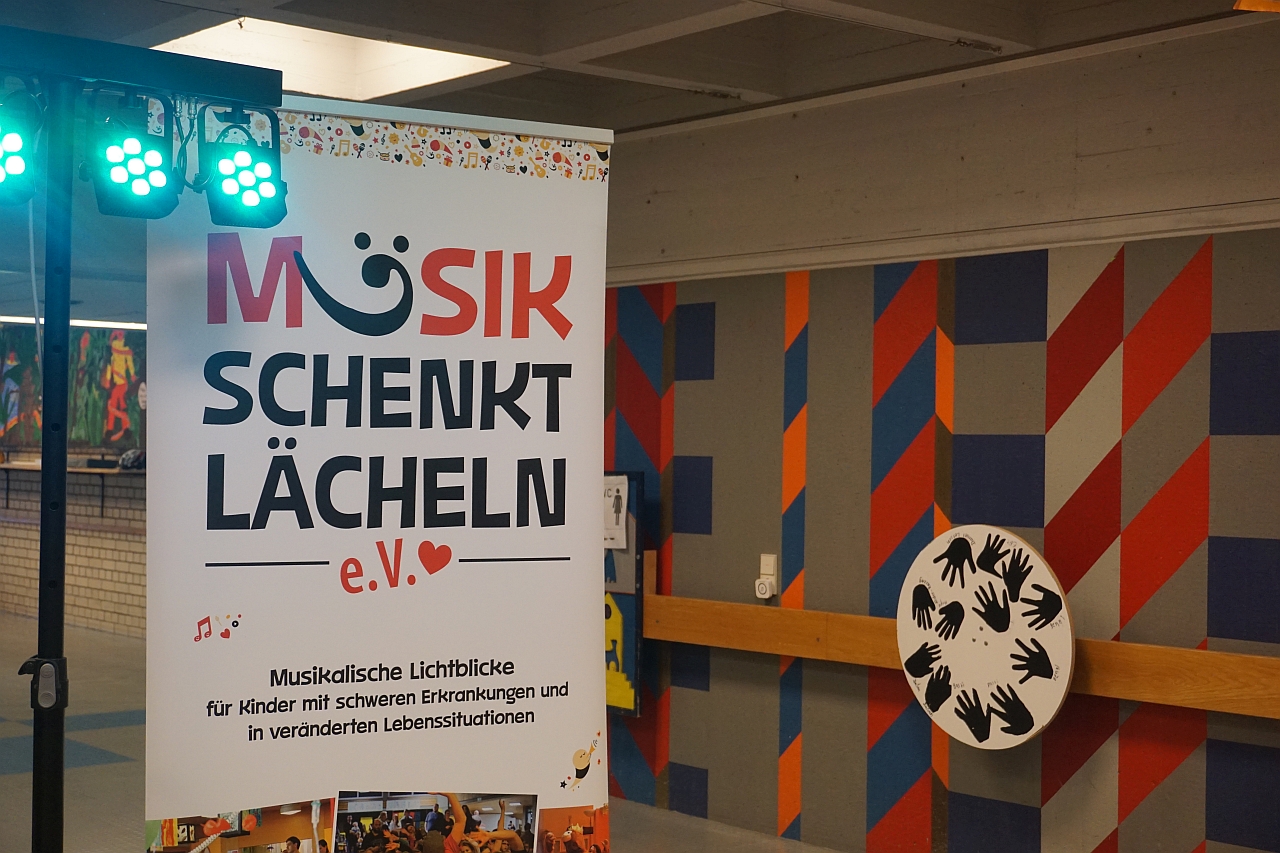 Plakat mit Text Musik schenkt Lächeln e.V.  Musikalische Lichtblicke für Kinder mit schweren Erkrankungen und in veränderten Lebenssituationen steht in der Aula neben der Bühne.