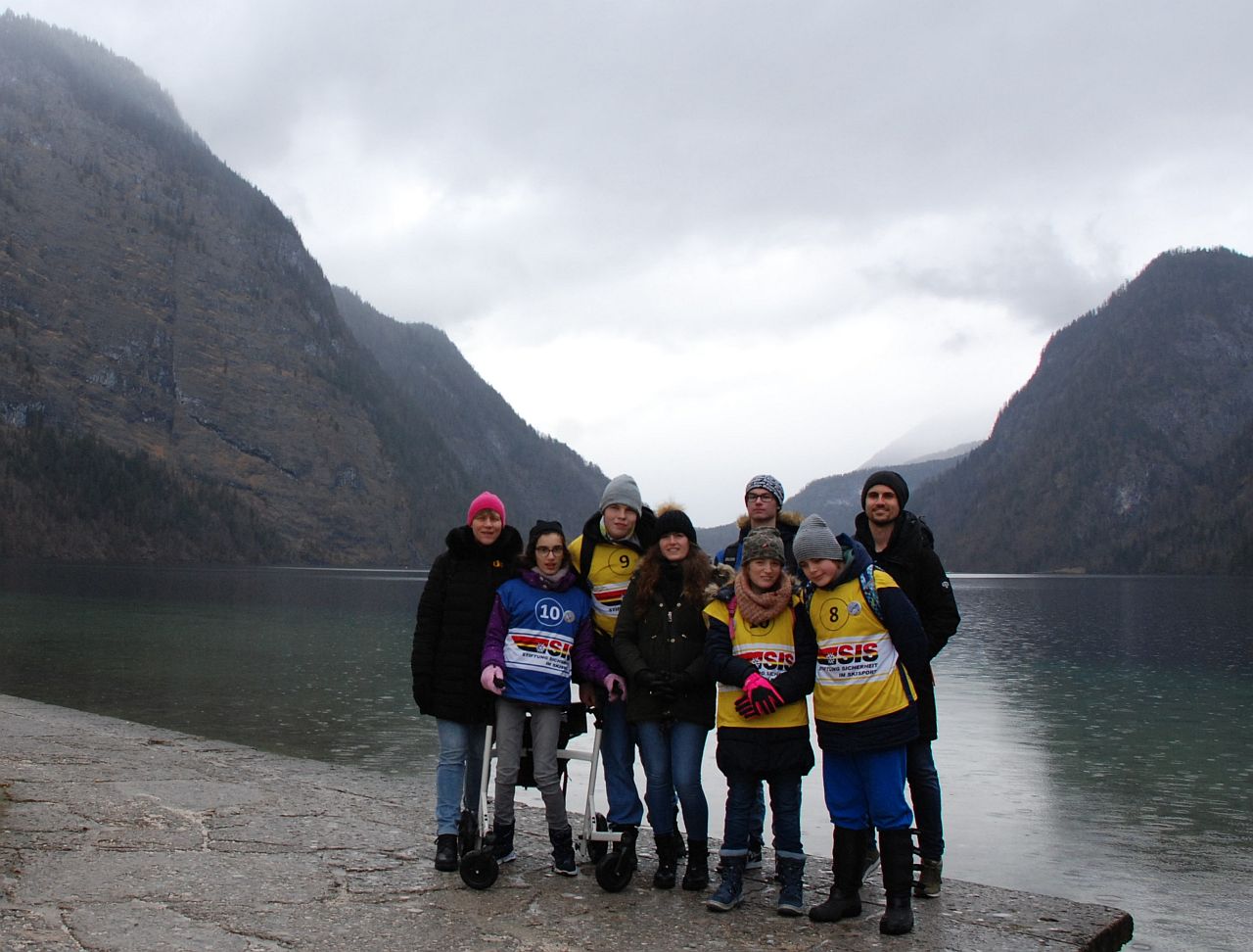 Frau Gauch, Marija, Julian, Frau Fecke, Florian, Lara, Christopher und Herr Kling stehen vor dem Königssee und lächeln in die Kamera. Hinter dem See sind wolkenverhangene Berge zu sehen.