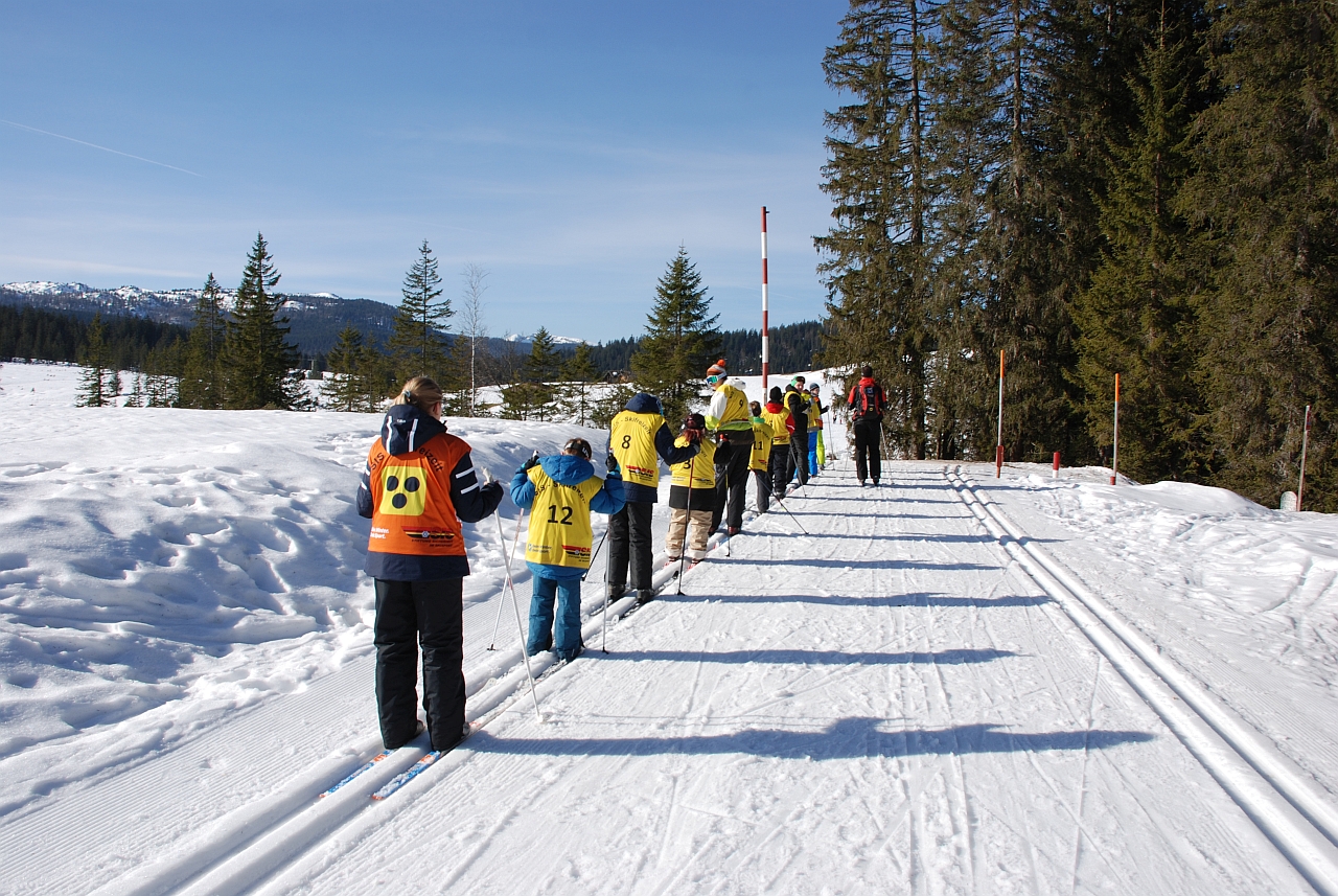 In einer Loipe stehen mehrere Kinder hintereinander. Alle tragen farbige Leibchen. Die Umgebung ist schneebedeckt. Rechts beginnt ein Wald. Im Hintergrund sind schneebedeckte Berge zu sehen.