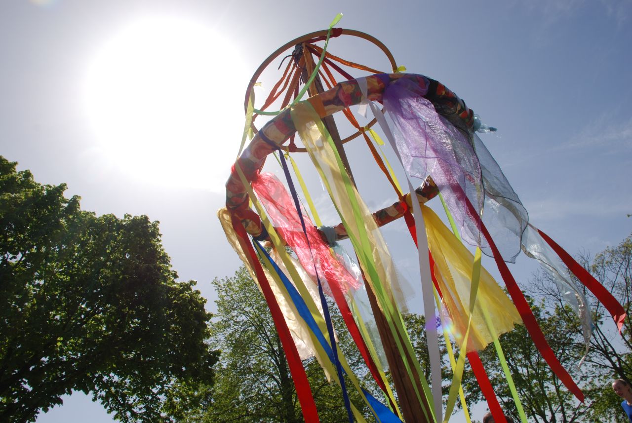 Gegen die strahlende Sonne sieht man die mit bunten Tüchern behangene Krone des Maibaums. Im Hintergrund sind grüne Bäume.