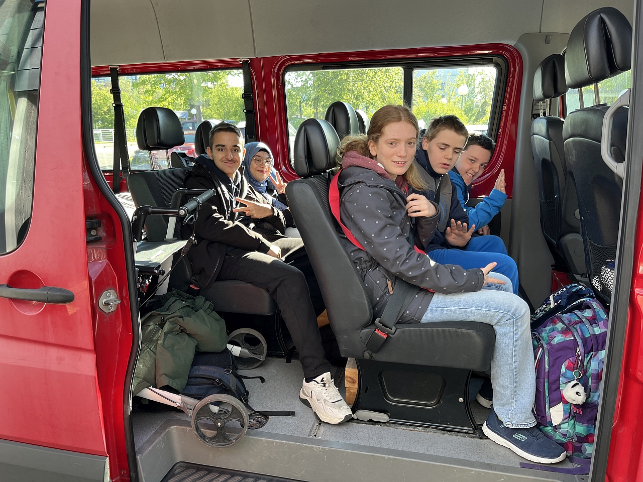 3 Jungs und 2 Mädchen in  Einem Bus und alle lachen. Im Bus sieht man auch Gepäck und einem Schulranzen und einen Rollator.