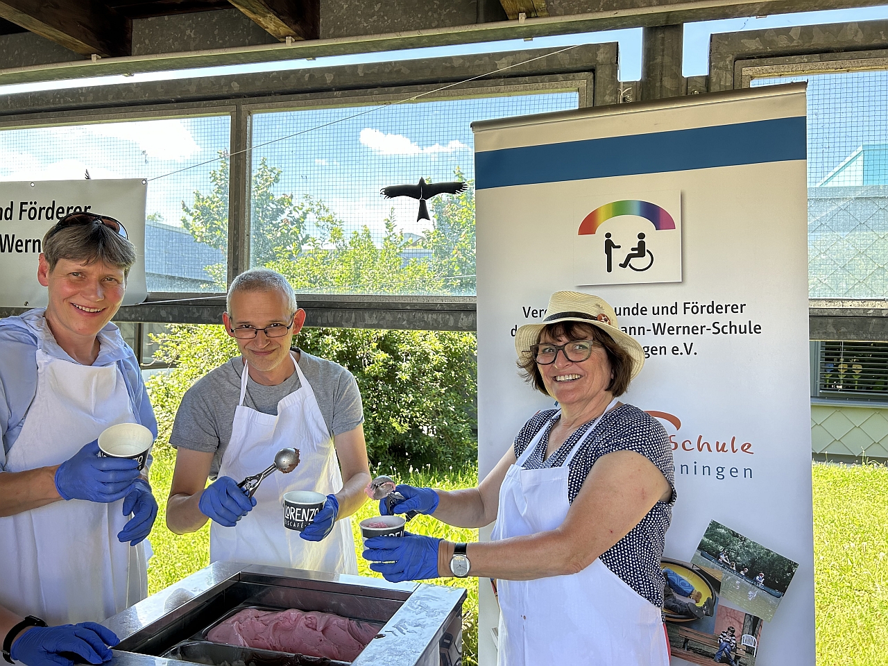 Drei Mitglieder des Vorstands stehen an der Eisausgabe. Sie tragen Schürzen und Handschuhe und haben Eislöffel und Eisbecher in der Hand. Im Hintergrund ist das Banner des Fördervereins zu sehen.
