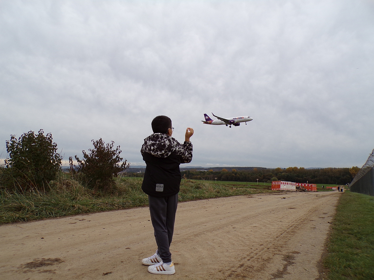 Ein Junge steht mit dem Rücken zur Kamera und schaut in den Himmel. Da ist ein großes Passagierflugzeug das gerade zur Landung ansetzt. Der Junge steht auf einem Feldweg. Der Himmel ist bewölkt.