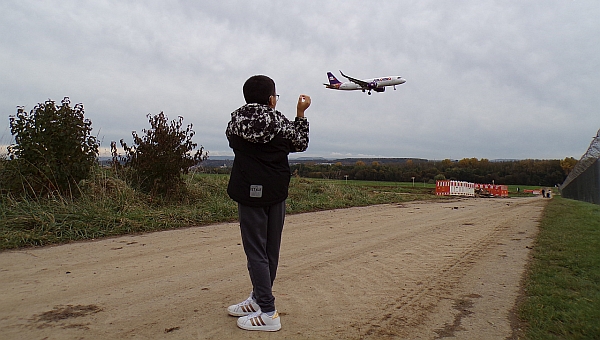 Ein Junge steht mit dem Rücken zur Kamera und schaut in den Himmel. Da ist ein großes Passagierflugzeug das gerade zur Landung ansetzt.  Der Junge steht auf einem Feldweg. Der Himmel ist bewölkt. 