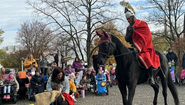 Ein als Sankt Martin verkleideter Reiter nähert sich auf einem schwarzen Pferd von rechts. In der Bildmitte sitzt ein als Bettler verkleideter Junge, neben ihm die Lehrerin. Schülerinnen und Schüler mit Laternen sitzen in Rollstühlen oder stehen mit Mitarbeitenden der Schule dahinter und schauen zu. 