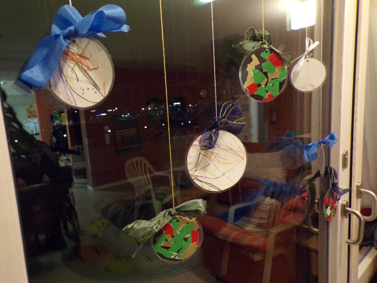 An einem Fenster hängen mehrere Kreise. Diese sind bemalt und dekoriert sodass sie wie Christbaumkugeln aussehen. Sie sind mit einem glitzernden Faden aufgehängt und haben auch eine Schleife
