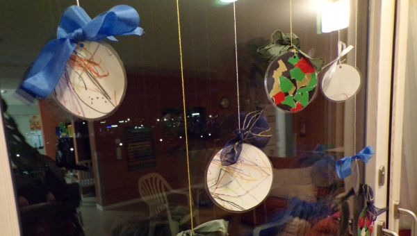  An einem Fenster hängen mehrere Kreise. Diese sind bemalt und dekoriert sodass sie wie Christbaumkugeln aussehen. Sie sind mit einem glitzernden Faden aufgehängt und haben auch eine Schleife