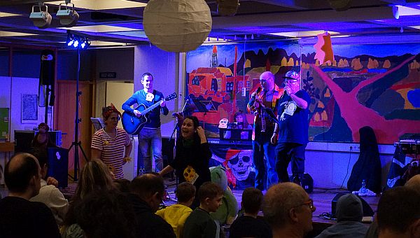 Vier Musiker sind auf der Bühne, links einer mit Gitarre, im Hintergrund eine Frau am Keyboard auf der rechten Seite zwei Sänger mit Mikrofonen. Zwei verkleidete Musikerinnen stehen vor der Bühne und animieren das Publikum 