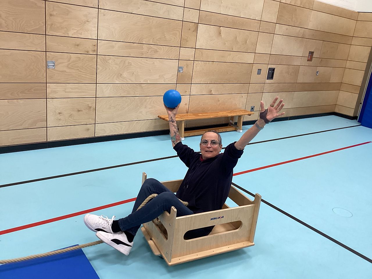 Eine Frau sitzt in einer Holzkiste auf Rollen. Diese steht in einer Turnhalle. Die Frau freut sich, sie hält ihre Arme in der Luft und hält einen blauen Ball in der Hand.