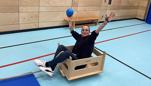 Eine Frau sitzt in einer Holzkiste auf Rollen. Diese steht in einer Turnhalle. Die Frau freut sich, sie hält ihre Arme in der Luft und hält einen blauen Ball in der Hand.