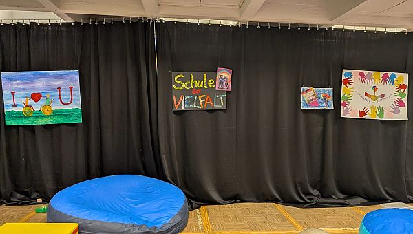 In der Aula hängen über der Bühne vier Plakate. Auf einem sieht man zwei Rollstuhlfahrende und die Botschaft „I love you“. Auf einem steht „Schule der Vielfalt“. Auf einem sind viele bunte Hände und darunter steht „Miteinander – Füreinander“.
