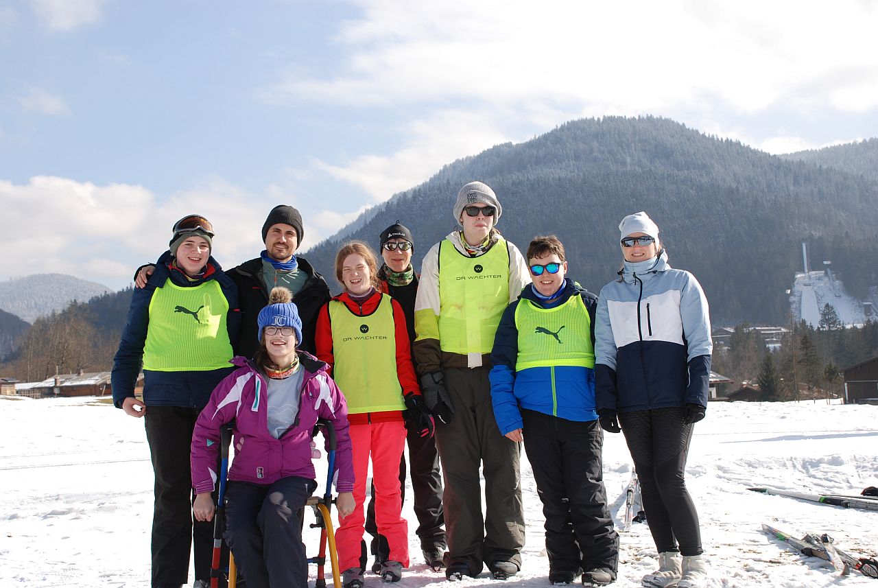 Vor einer traumhaft schönen Schneelandschaft mit blauem Himmel und ein paar wenigen Wolken stehen von links nach rechts Christopher, Marija, Herr Kling, Lara, Frau Gauch, Julian, Josua und Frau Fecke. Alle lächeln in die Kamera.