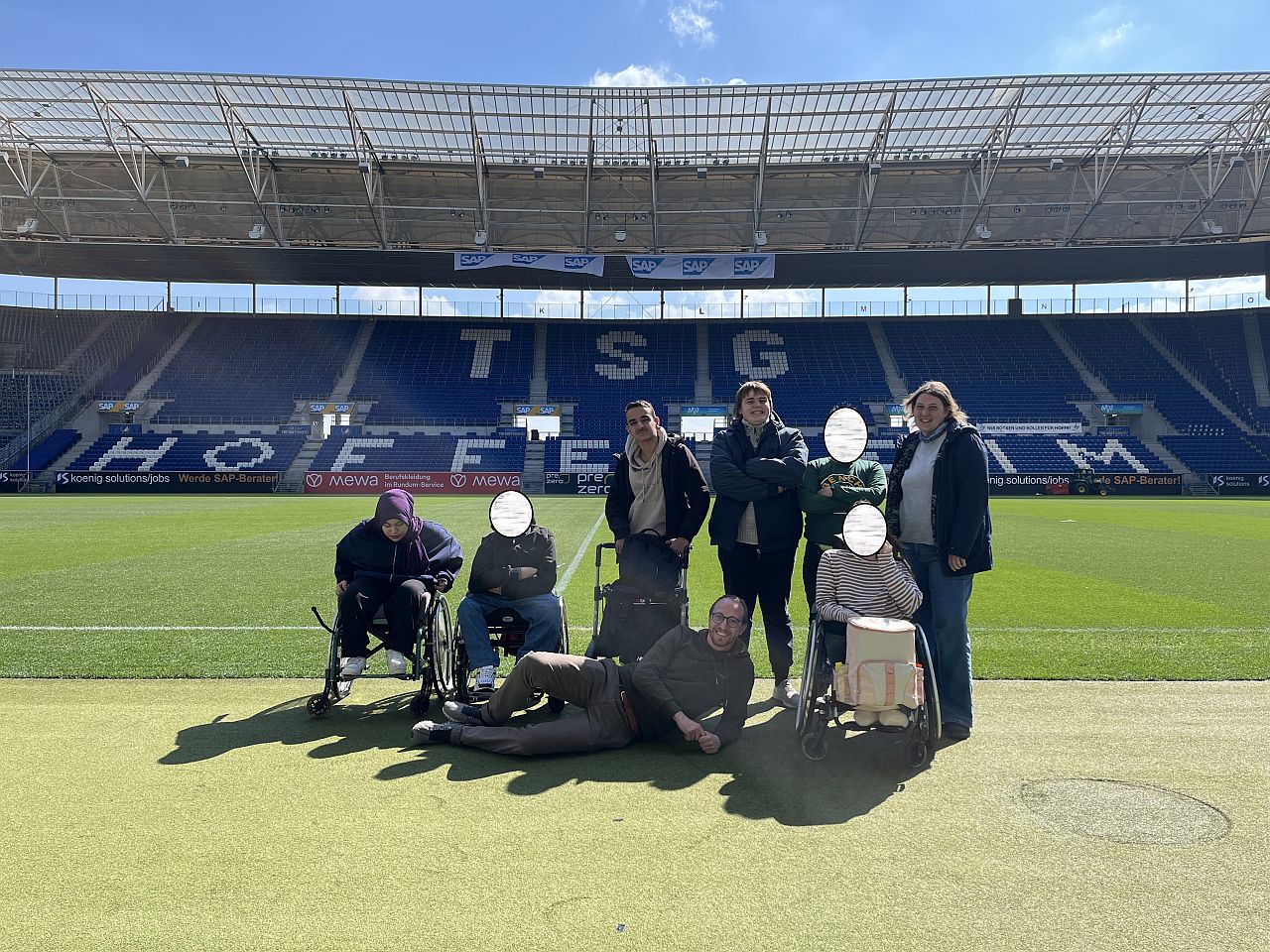 Sechs Kinder und zwei Lehrer machen ein Foto im Fußballstadion. Das Wetter ist sonnig und der Lehrer liegt auf dem Boden vor den Schülern und schaut in die Kamera.