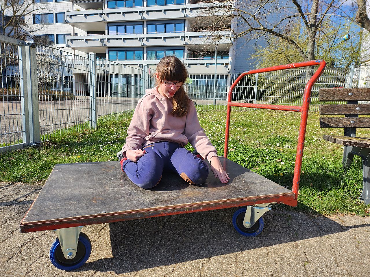 Schülerin sitzt auf dem Transportwagen und lacht. Im Hintergrund ist das Internat zu sehen