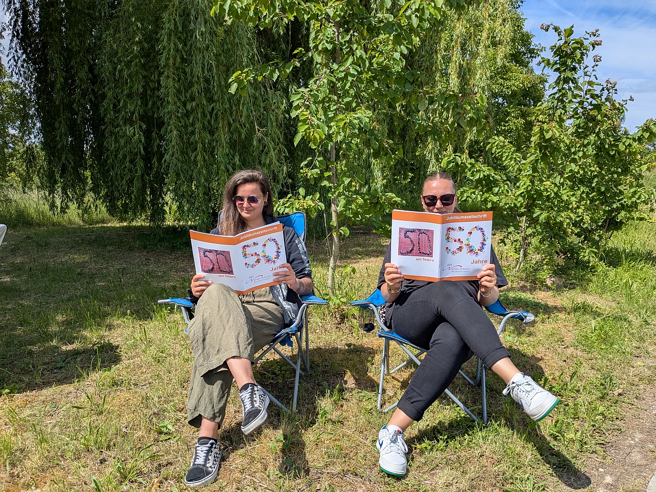 2 Frauen sitzen mit der Festtagszeitung in Campingstühlen und lesen diese durch.