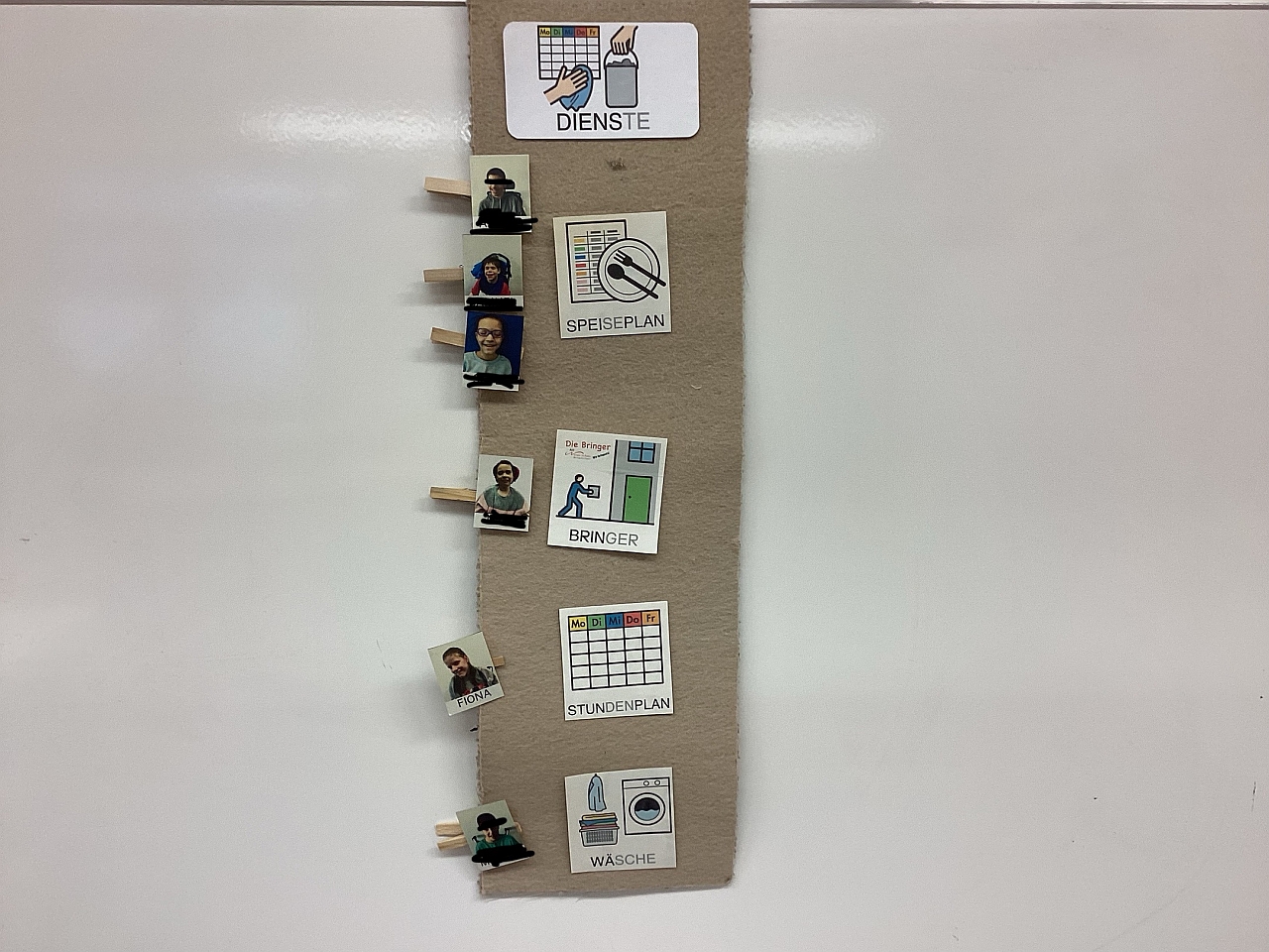 Auf einer Tafel hängen unter der Überschrift Dienste vier Bildkarten: Speiseplan, Bringer, Wäsche, Stundenplan. Zu jeder Bildkarte hängen ein odere mehrer Fotos eines Schülers oder einer Schülerin. 