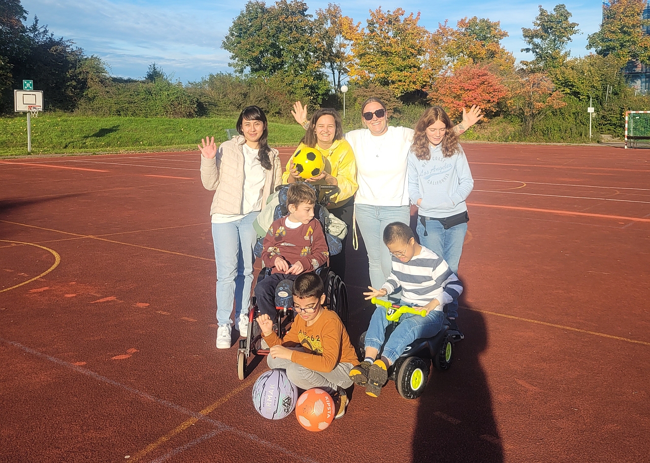Auf dem Foto sieht man eine Gruppe auf einem Sportplatz. Hinten stehen drei Erwachsene und ein junges Mädchen. In der Mitte ist ein Junge im Rollstuhl und ein anderer auf einem Bobbycar. Vorne kniet ein Junge. Er hat zwei Bälle in den Händen. Alle lachen. Die Sonne scheint.