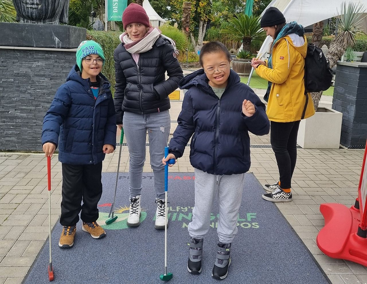 Drei Kinder sind zu sehen: zwei Jungen und ein Mädchen. Sie stehen nebeneinander. Das Mädchen steht in der Mitte.Sie sind draußen. Alle tragen Jacken und Mützen.Jedes Kind hält einen Minigolf-Schläger in der Hand.