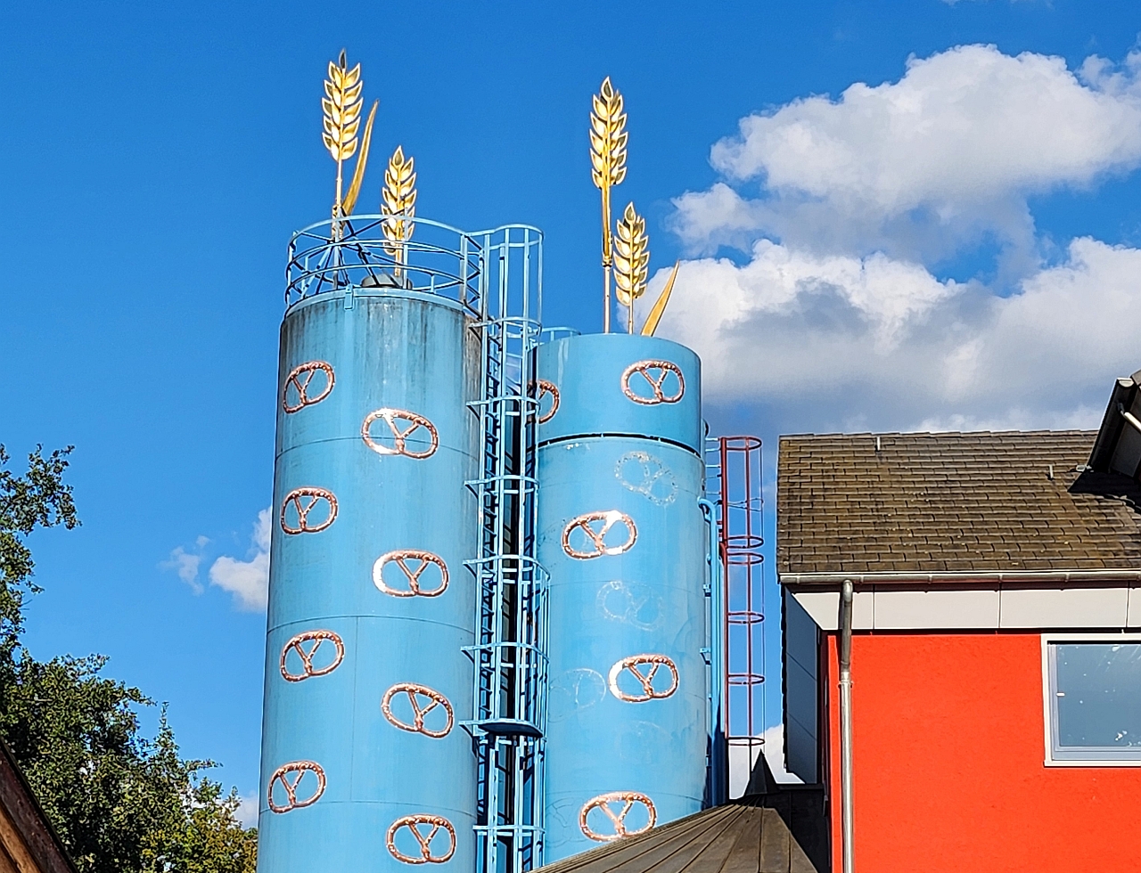 Der Himmel ist ein wunderschöner Blauton, zwei weise Wolken im hintergrund, zwei Blaue türme mit gelben Brezeln, auf den Türmen sind zwei Kornblumen zu sehen. 