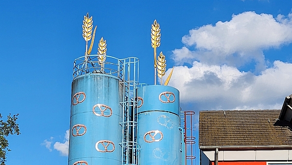 Der Himmel ist ein wunderschöner Blauton, zwei weise Wolken im hintergrund, zwei Blaue türme mit gelben Brezeln, auf den Türmen sind zwei Kornblumen zu sehen. 