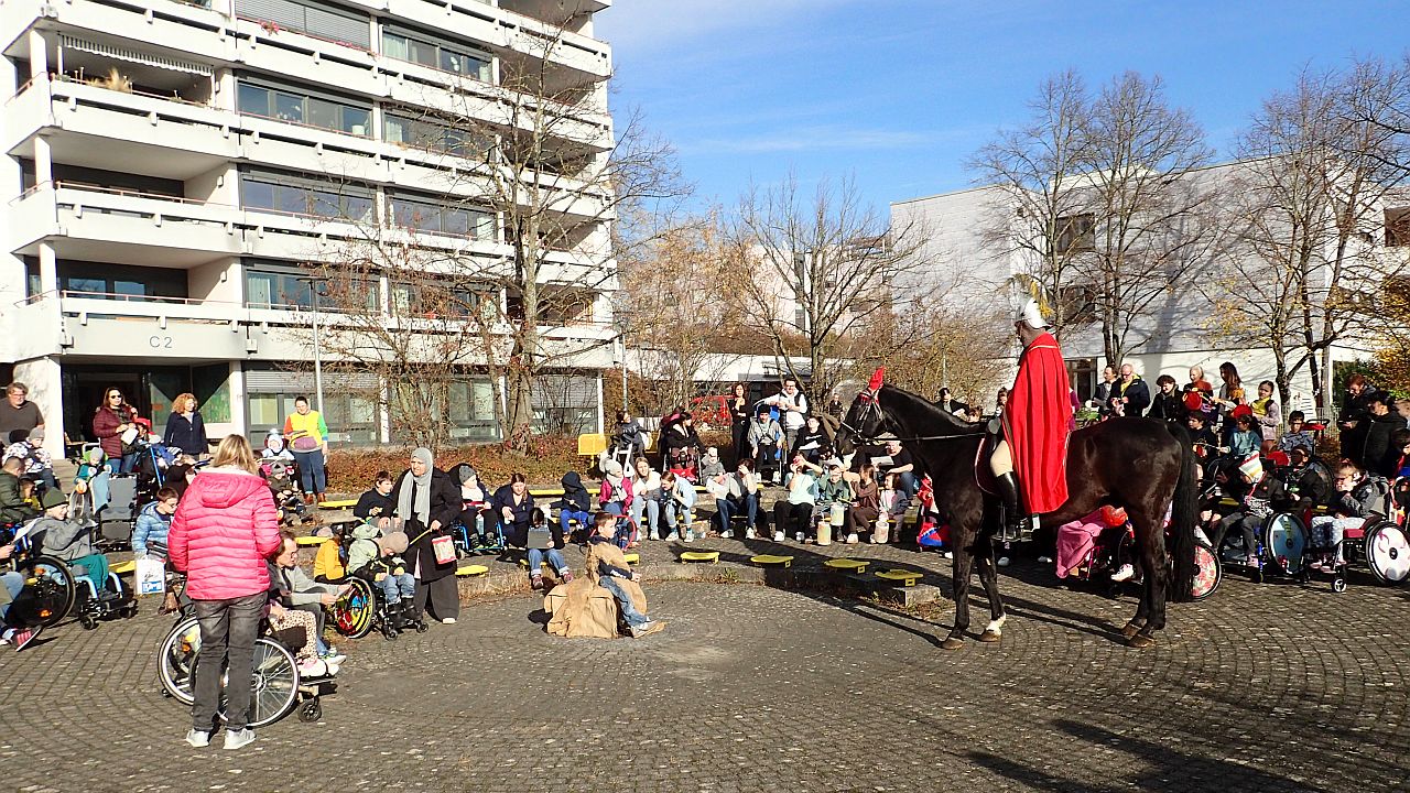 Runder Schulhof, im Hintergrund mehrstöckiges Internats- und Schulgebäude. Ringsum sitzen Kinder, Jugendliche und Erwachsene, zum Teil im Rollstuhl, manche mit Laternen. Mittig ein Junge, als Bettler verkleidet, von rechts kommt Sankt Martin auf dem Pferd im roten Mantel.