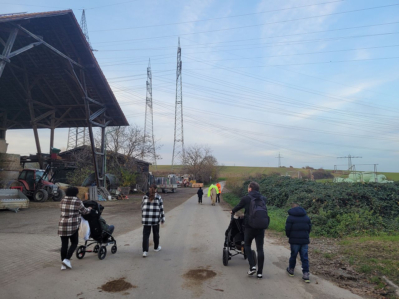 Man sieht eine Gruppe von 6 Leuten von hinten. Zwei davon schieben einen Buggy. Ganz vorne läuft ein Pferd an einer Leine. Die Leute sind auf einem Bauernhof