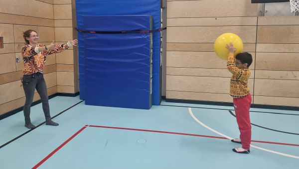 Eine Frau und ein Junge stehen sich gegenüber. Sie spielen sich einen gelben Ball zu. Sie sind in einer Sporthalle