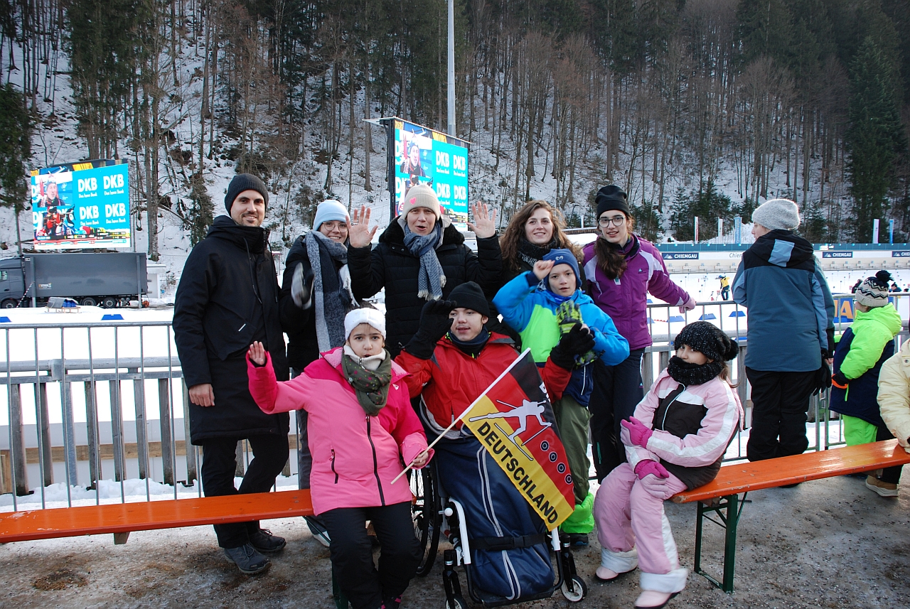 Vor einem Geländer stehen Herr Kling, Lena, Frau Gauch, Frau Fecke, Marija, Ghofran, Maik, Luca und Sophia. Hinter dem Geländer sieht man Schnee und die Schießanlage vom Biathlon. Dahinter geht es einen Hang hinauf, auf dem Bäume wachsen.
