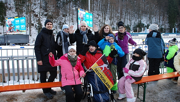 Vor einem Geländer stehen Herr Kling, Lena, Frau Gauch, Frau Fecke, Marija, Ghofran, Maik, Luca und Sophia. Hinter dem Geländer sieht man Schnee und die Schießanlage vom Biathlon. Dahinter geht es einen Hang hinauf, auf dem Bäume wachsen.