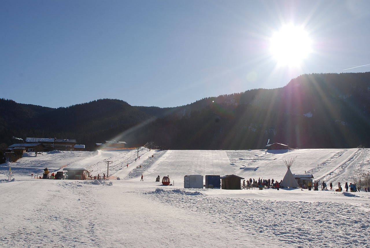 An einem sonnenbeschienenen Schneehang sind drei verschiedene Schlepplifte zu sehen. Unten sind einige Hütten sowie ein Tipi und es tummeln sich viele Menschen. Oberhalb des Hangs ist ein bewaldeter Berg zu sehen. Darüber blauer Himmel und strahlende Sonne.