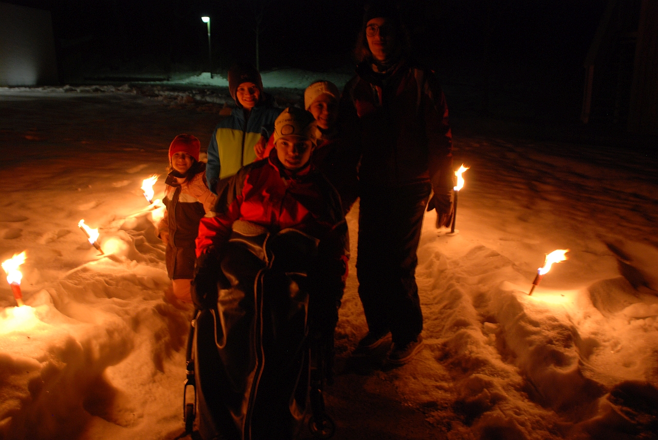 Sophia, Luca, Ghofran, Maik und Marija stehen auf einem Weg, der rechts und links von brennenden Fackeln gesäumt ist, die im Schnee stecken. Alle lächeln in die Kamera.