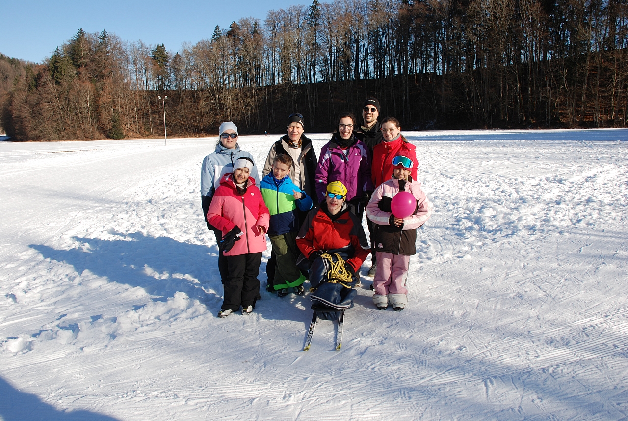 Im Schnee stehen Frau Fecke, Frau Gauch, Marija, Herr Kling, Lena, Ghofran, Luca, Maik und Sophia und lächeln in die Kamera. Im Hintergrund sieht man die Loipen, dunkle Bäume und blauen Himmel.