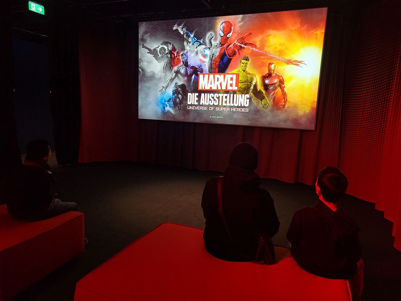 Man sieht eine Leinwand auf der verschiedene Superhelden zu sehen sind. In der Mitte steht der Schriftzug Marvel 