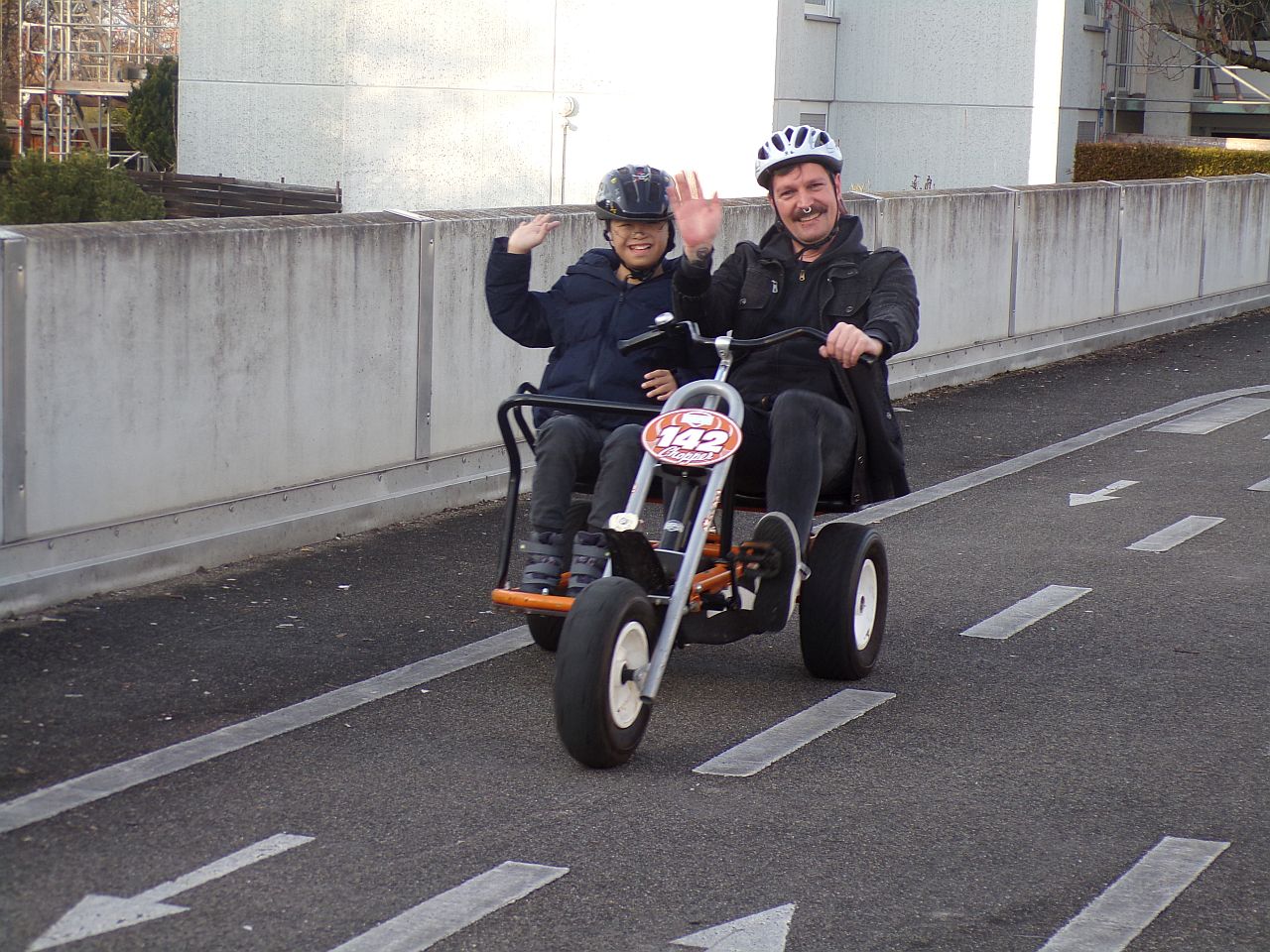 Ein Junge und ein Mann sitzen in einem Kettcar und winken in die Kamera.