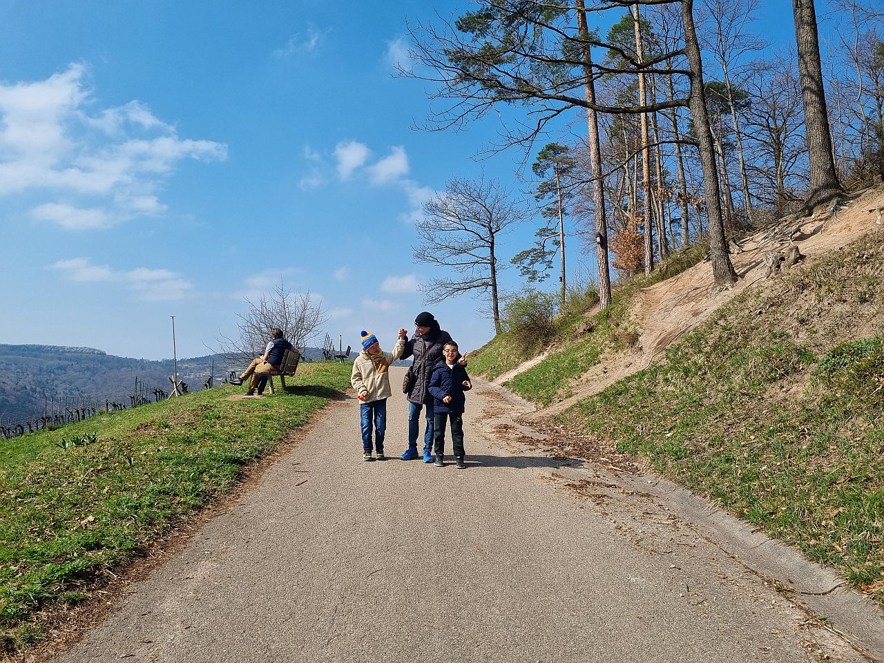 Eine Frau steht mit zwei Jungen auf einem Weg. Rechts neben dem Weg stehen Bäume und links ist eine Wiese. Die Sonne scheint. 