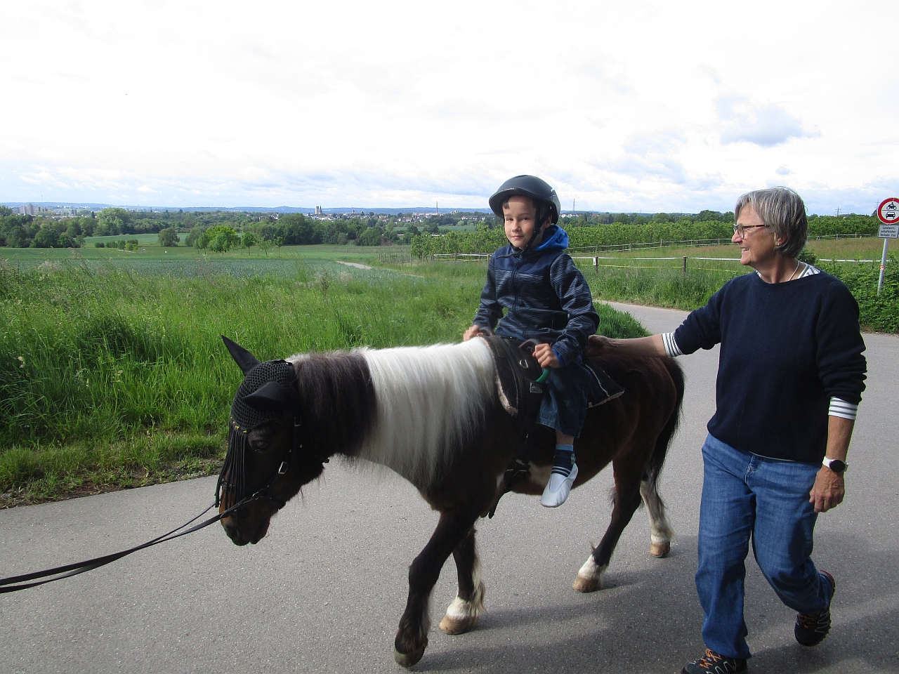 Ein neunjähriger Junge auf einem dunkelbraun-weiß gefleckten Pony, das nach links läft. Links im Bild die Zügel, rechts neben dem Jungen eine Frau, die ihn begleitet. Im Hintergrund Felder am Rand der Karlshöhe Ludwigsburg.