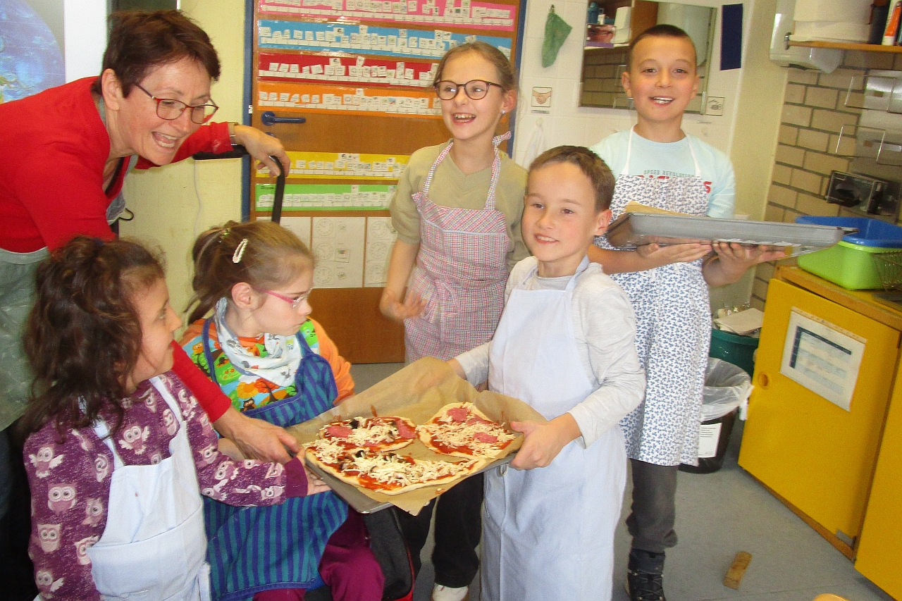 2 Jungen und 3 Mädchen im Grundschulalter, davon ein Mädchen im Rollstuhl, präsentieren mit ihrer Lehrerin selbst belegte Pizza auf 2 Blechen