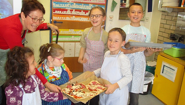 2 Jungen und 3 Mädchen im Grundschulalter, davon ein Mädchen im Rollstuhl, präsentieren mit ihrer Lehrerin selbst belegte Pizza auf 2 Blechen