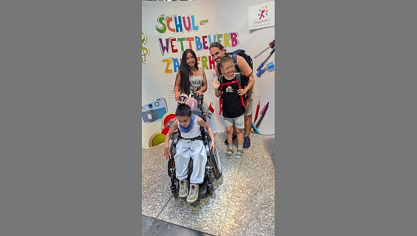 Fernanda, Herr Kling, Muratcan und Henry stehen vor einem Plakat mit der Aufschrift „Schulwettbewerb Zauberhaft“ und lächeln in die Kamera.