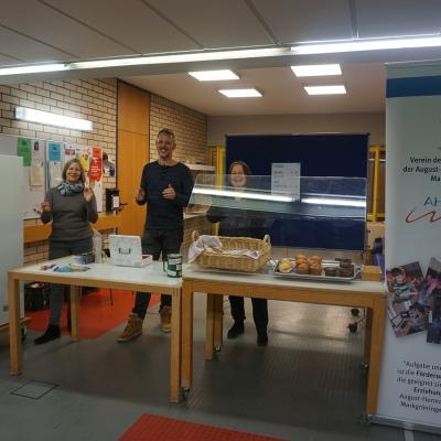 Plakat des Fördervereins rechts neben dem Stand mit der Bewirtung. Drei Mitarbeiter die hinter den Tischen stehen. Auf den Tischen sind die Kasse, ein Korb mit Brezeln und eine Platte mit Muffins. Verschiedene Getränke im Kühlschrank links neben der Theke