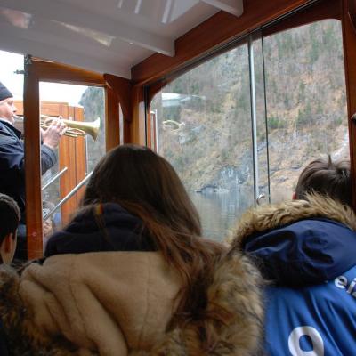 Der Bootsführer steht an der Tür des Boots und spielt mit einer Trompete in Richtung Felswand. Frau Fecke und Florian schauen gebannt zu.