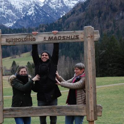 Vor einem schönen Bergpanorama steht ein hölzerner Rahmen. Darauf steht „Ruhpolding Madshus“. Herr Kling hängt am oberen Rand des Rahmens. Frau Fecke und Frau Gauch kitzeln ihn von den Seiten.