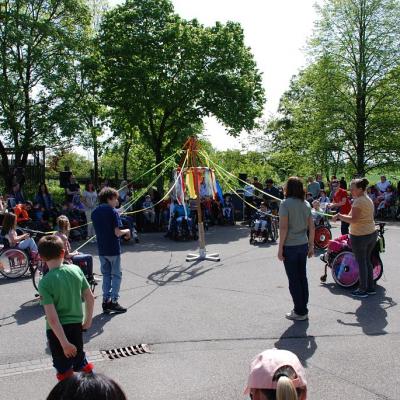 Die Tanz-AG tanzt um den Maibaum. Sie halten lange bunte Bändel, die an der Spitze des Baums befestigt sind. Darum herum stehen und sitzen die Mitglieder der Schulgemeinschaft.