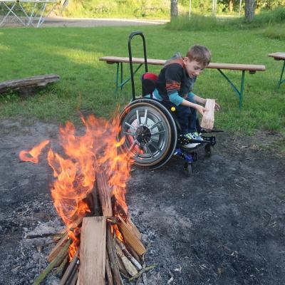 Hinter dem Lagerfeuer lädt ein Schüler ein Holzscheit auf seinen Rollstuhl