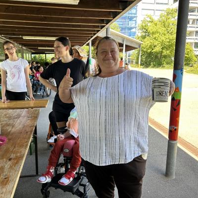 Eine Mitarbeiterin hat viele leere Eisbecher in der Hand und zeigt mit den Daumen hoch, Im Hintergrund sind Kinder und Erwachsene beim Eis essen 