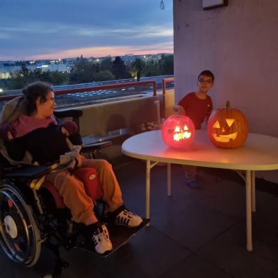 Ein Mädchen im Rollstuhl und ein Junge stehen auf einem Balkon. Es ist dunkel. Vor dem Jungen steht ein Tisch. Auf diesem sind zwei Kürbisse. Die Kürbisse haben eingeschnitzte Gesichter. Aus denen Leuchtet es hell. 