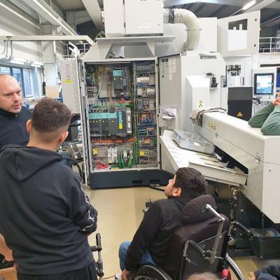 Links ist ein Mann von Spinner Automation. Vor ihm sind vier Schüler: innen. Der Mann erzählt den Schüler: innen etwas über den Betrieb. Im Hintergrund steht eine große Maschine.