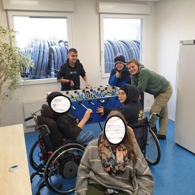 Sechs Schüler: innen verteilen sich um einen Tischkicker und lächeln in die Kamera. Der Tischkicker steht in einem Pausenraum.